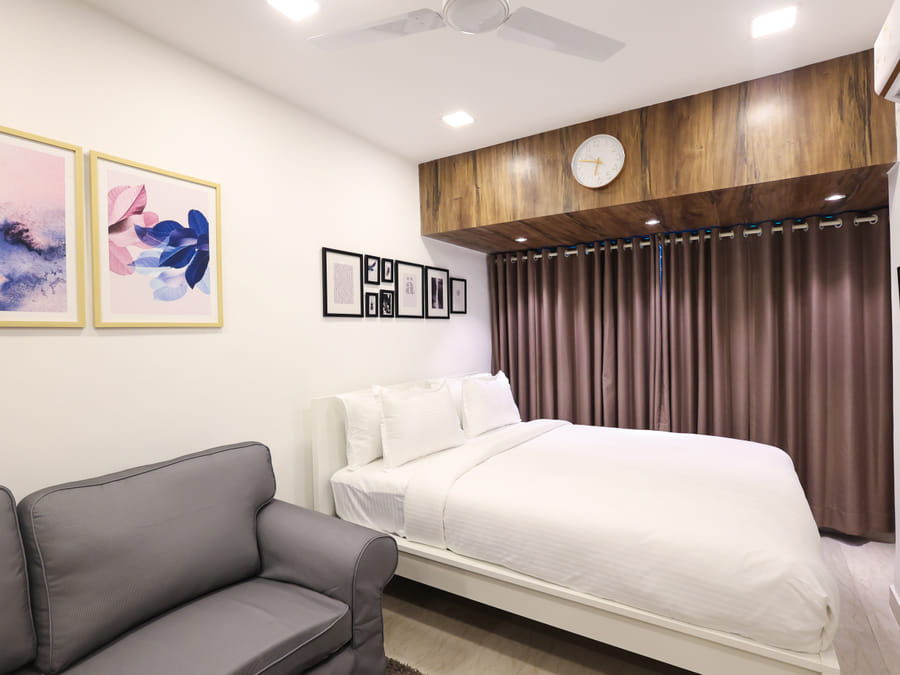Bedroom of Rooms 402, 3001, and 5001 at Wabi-Sabi, Juhu, with a wooden panel clock above the bed, a grey sofa, and colourful framed floral art.