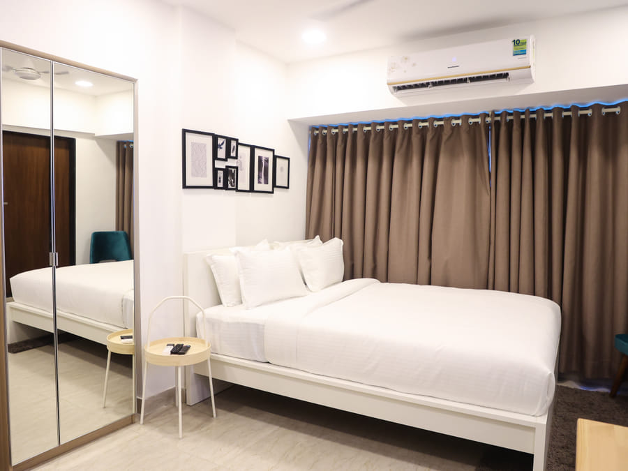 Bedroom of Rooms 403, 3003, and 5003 at Wabi-Sabi, Juhu, showing a full-length mirrored wardrobe beside the bed, with the teal chair reflected in it.
