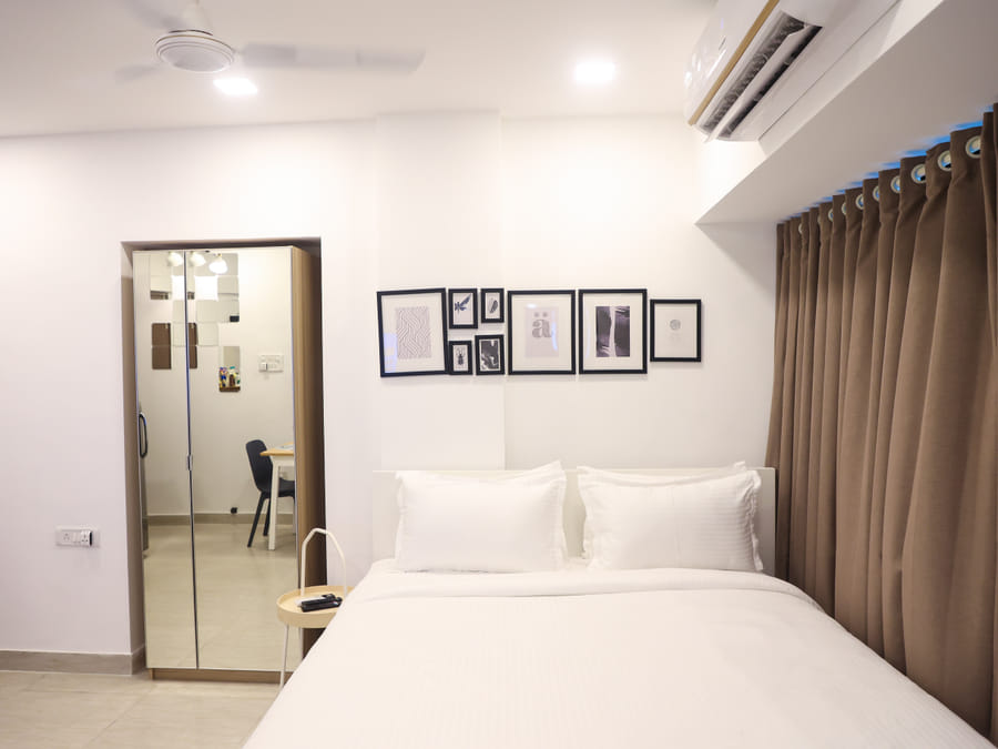 Bedroom of Rooms 403, 3003, and 5003 at Wabi-Sabi, Juhu, with brown curtains, a round side table with remote, and a mirrored wardrobe by a desk area.