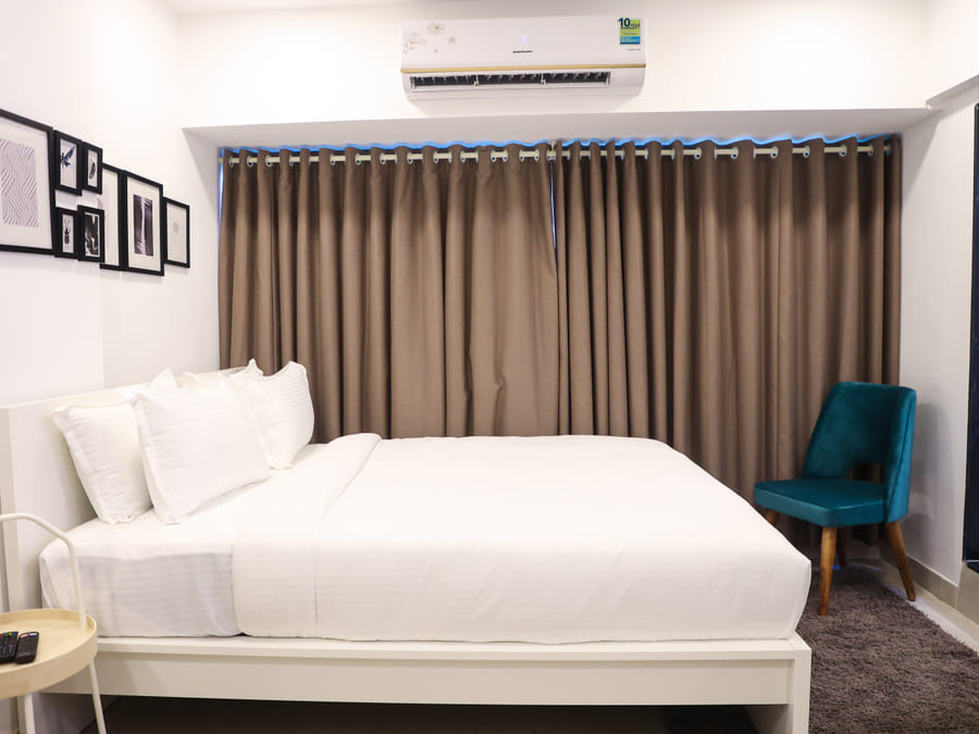 Bedroom of Rooms 403, 3003, and 5003 at Wabi-Sabi, Juhu, with a teal velvet accent chair beside brown curtains and a dark grey rug at the foot of the bed.