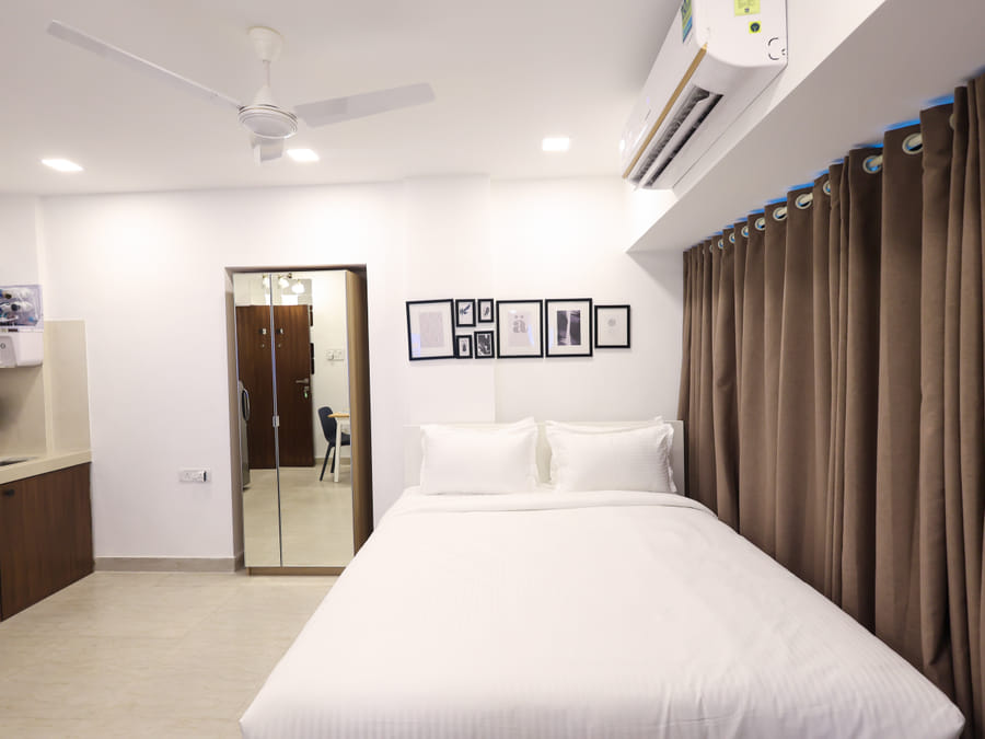Bedroom of Rooms 403, 3003, and 5003 at Wabi-Sabi, Juhu, with floor-to-ceiling brown curtains and an open kitchenette visible beyond the bed.