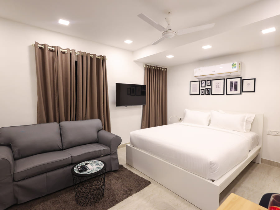 Studio layout of Rooms 404, 3003, and 5003 at Wabi-Sabi, Juhu, with a grey sofa and coffee table on a rug, a wall-mounted TV, and the bed beyond.