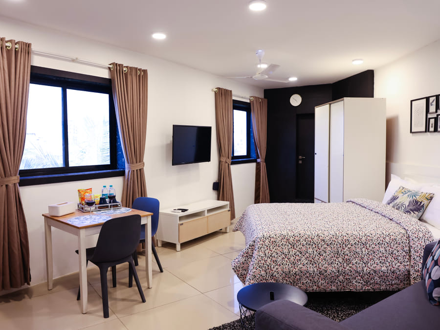Bedroom of Room 501 at Wabi-Sabi, Juhu, with a patterned bedspread, wall-mounted TV, dining table with 2 chairs, a white wardrobe, and curtained windows.