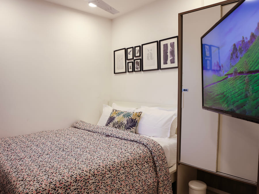 Bedroom of Room 502 at Wabi-Sabi, Juhu, closer view of the leaf-print cushion and headboard beside a wardrobe with an arrow decal and a TV showing tea fields.