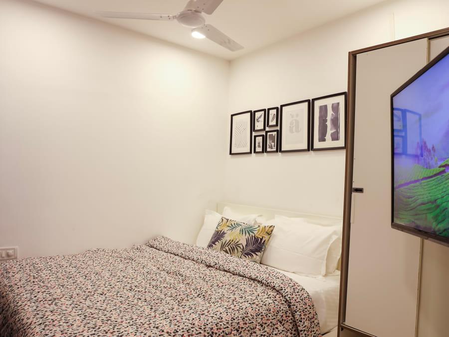 Bedroom of Room 502 at Wabi-Sabi, Juhu, with a printed duvet, a leaf-print accent cushion, a wall-mounted TV showing a green landscape, and a white wardrobe.