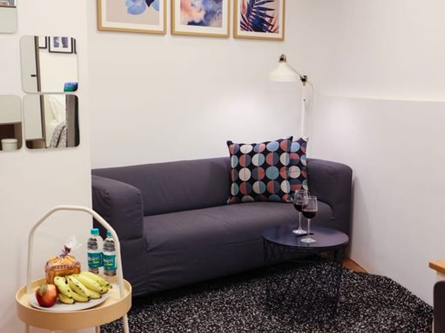 Living area at Wabi-Sabi, Juhu, with a navy sofa with printed cushions, a wall-mounted lamp, 3 framed art prints, a mirror mosaic, and a side table with fruit.