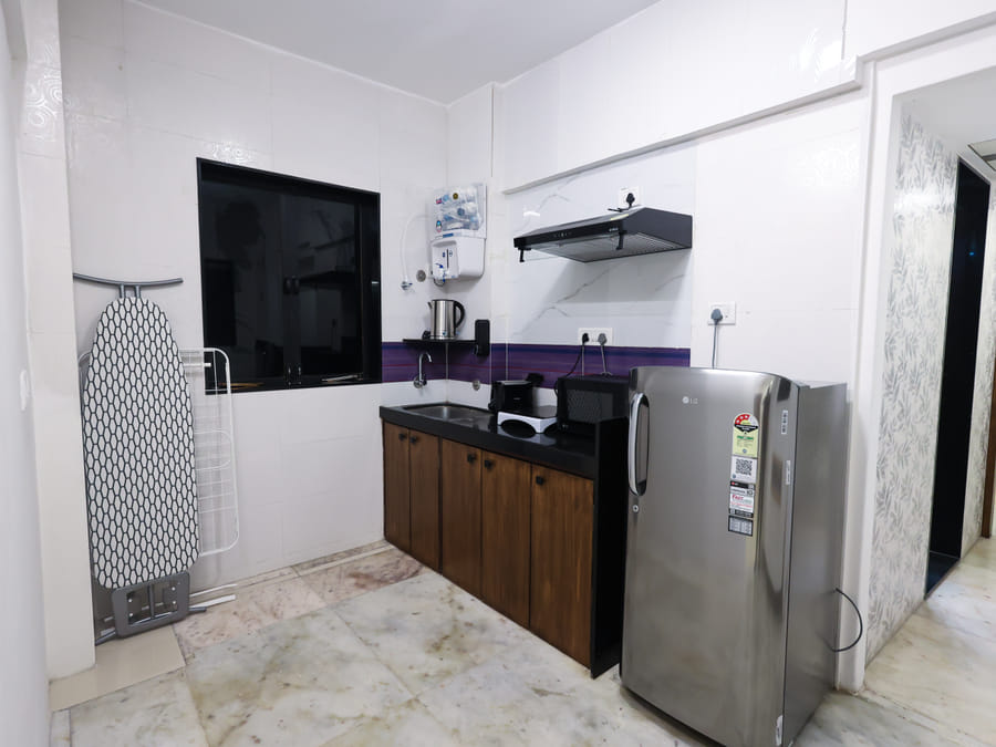 Kitchen in Room 401 at Wabi-Sabi, Juhu, with a black countertop, purple-striped backsplash, sink, exhaust hood, fridge, an ironing board, and a window.