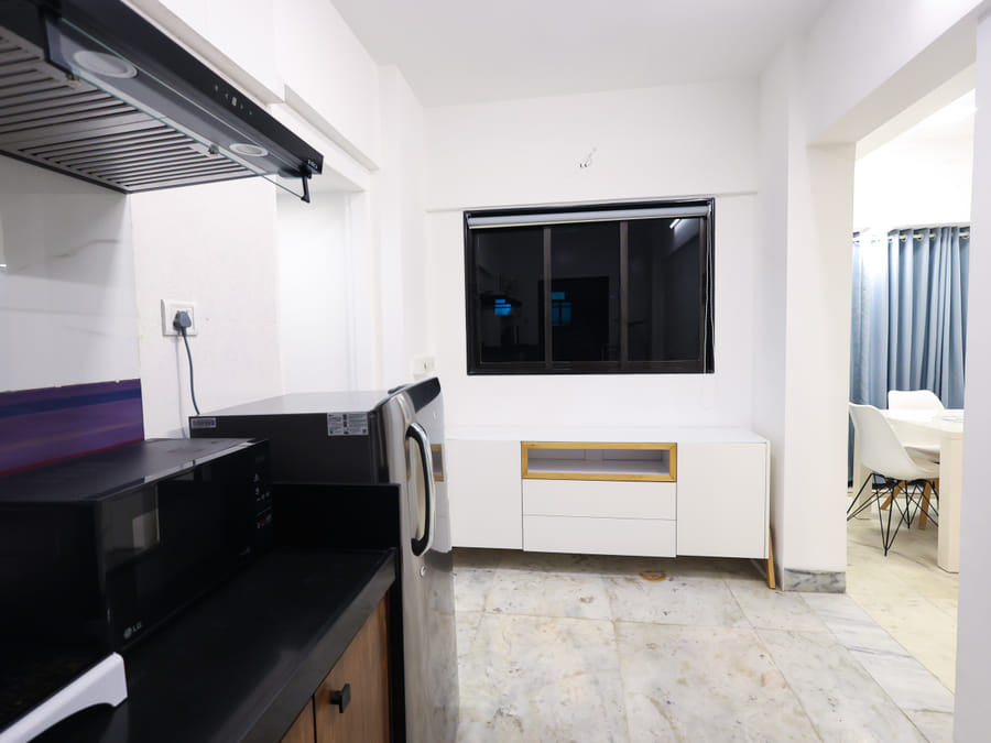 Kitchen in Room 401 at Wabi-Sabi, Juhu, with a fridge and black countertop beside a white TV console, opening onto a dining area with blue curtains.