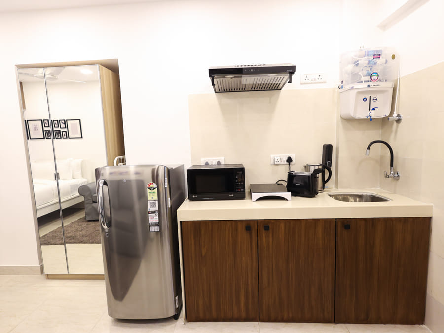 Kitchenette at Wabi-Sabi, Juhu, with a fridge, microwave, toaster, sink, and a mirrored wardrobe reflecting the bedroom with framed wall art.