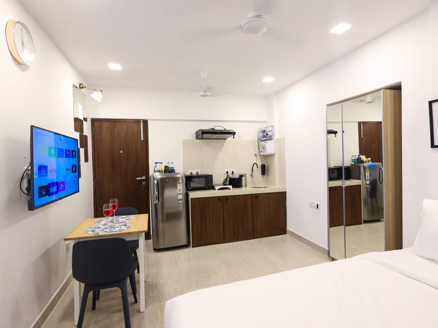 Studio room at Wabi-Sabi, Juhu, showing a bed, mirrored wardrobe, wall-mounted TV, dining table with 2 chairs, and a kitchenette with fridge and microwave.
