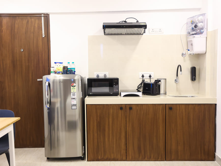 Kitchenette at Wabi-Sabi, Juhu, with an LG fridge, microwave, toaster, kettle, exhaust hood, sink, and a wall-mounted water purifier over wooden cabinets.