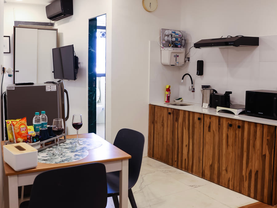 Open-plan kitchen and dining area in Room 501 at Wabi-Sabi, Juhu, with a wooden dining table, 2 black chairs, wine glasses, a wardrobe, TV, and fridge.