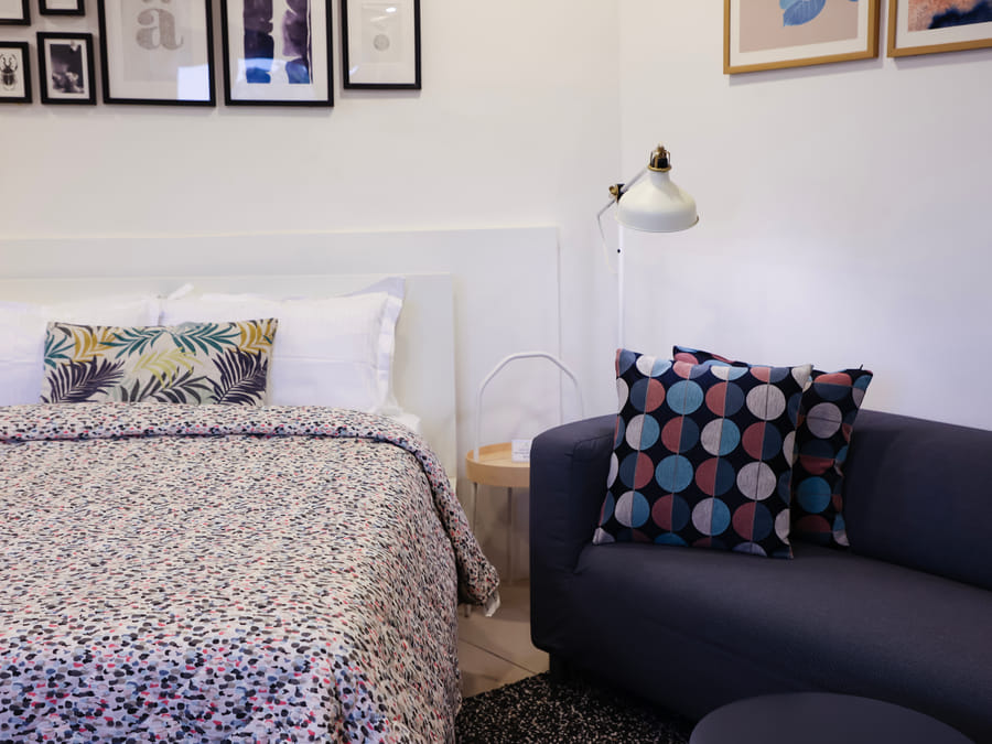Bed with a patterned bedspread and a navy sofa with printed cushions in Room 501 at Wabi-Sabi, Juhu, with framed wall art and a wall-mounted reading lamp.