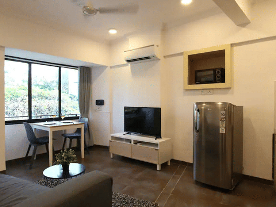 Living area at Wabi-Sabi, Juhu, with a grey sofa, coffee table, TV console, wall-mounted AC, fridge, and a window overlooking greenery beside a dining table.