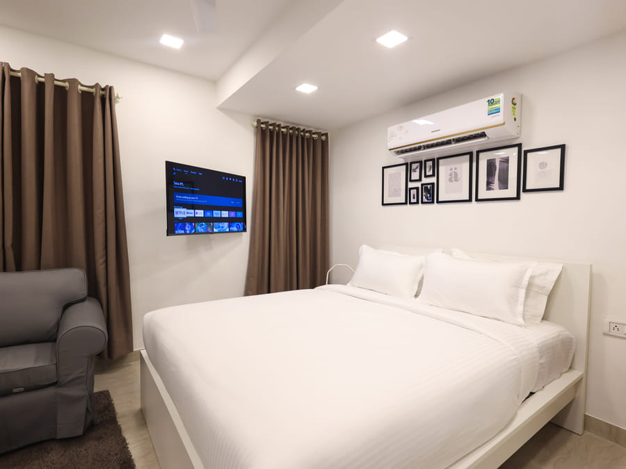 Bedroom at Wabi-Sabi, Juhu, with a grey armchair, brown curtains, a wall-mounted smart TV on its setup screen, and a gallery wall of framed art.