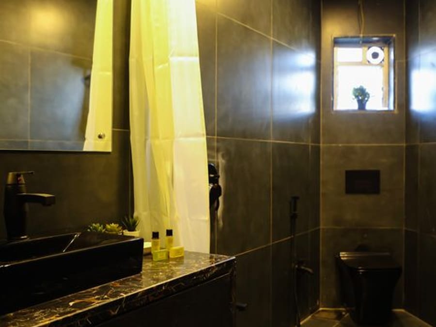 Bathroom at Wabi-Sabi, Juhu, with dark tiled walls, a black stone washbasin, a mirror, a shower curtain, a black toilet, and a small frosted window.
