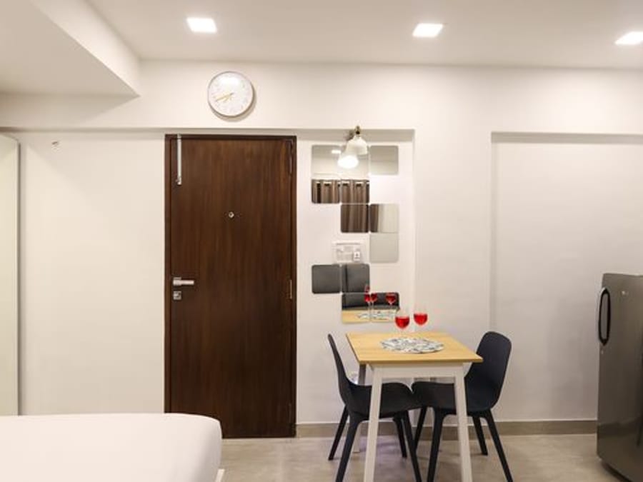 Studio room at Wabi-Sabi, Juhu, with a wooden entry door beneath a wall clock, a dining table with wine glasses, and a refrigerator by the kitchenette.