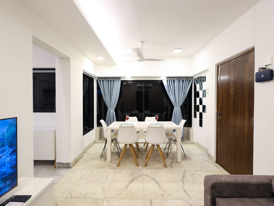 An open dining and living area at Wabi-Sabi, Juhu with a marble floor, a white dining table with 6 chairs, a television on a low console, and a grey sofa.