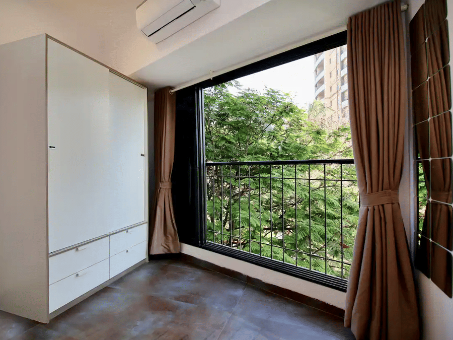 Room in a service apartment with a white sliding-door wardrobe, wall-mounted AC unit, and a large window with brown curtains overlooking trees and a building.