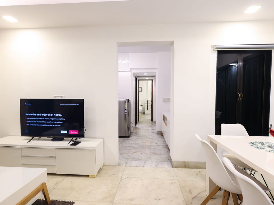 Living area at Wabi-Sabi, Juhu, with a marble floor, a TV on the Netflix sign-up screen, a dining table, and a hallway leading to the bedroom.