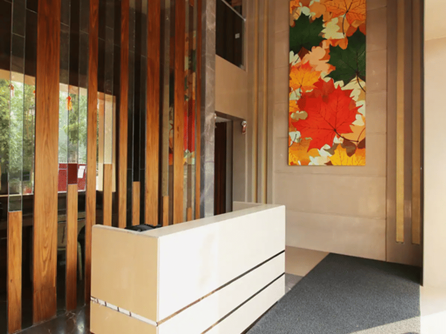 The reception lobby at Wabi-Sabi, Juhu with a white front desk, wooden slat partition, a large autumn-leaf wall art, and a grey carpeted floor.