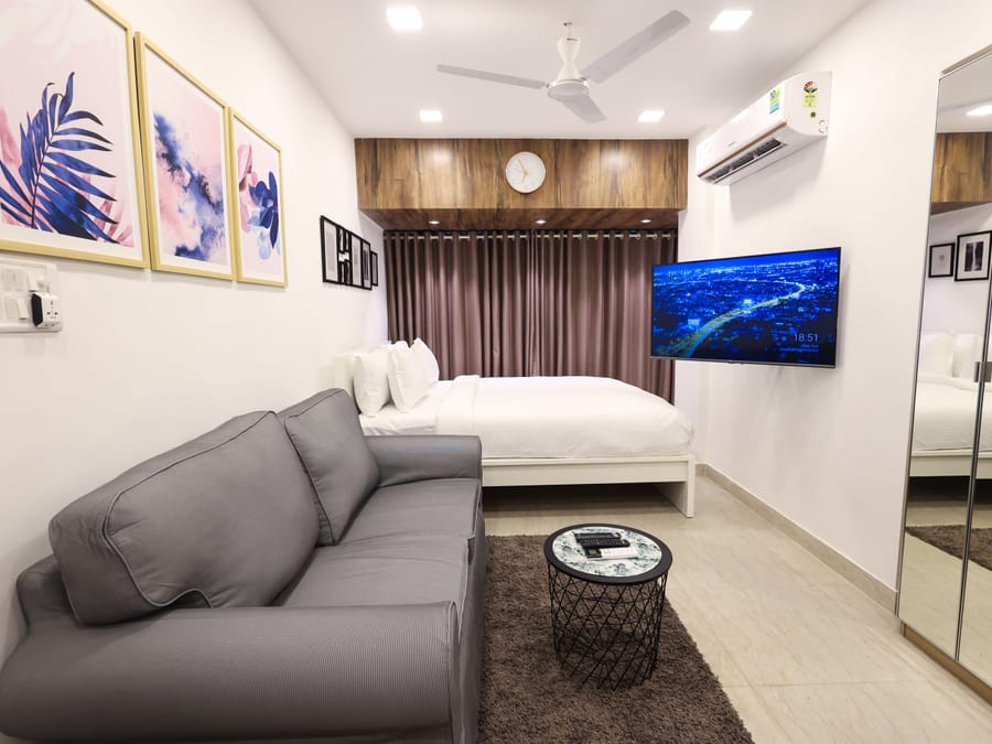 A fully furnished studio apartment at Wabi-Sabi, Juhu with a grey sofa, white bed, wall-mounted television, mirrored wardrobe, and a wooden headboard with a clock.
