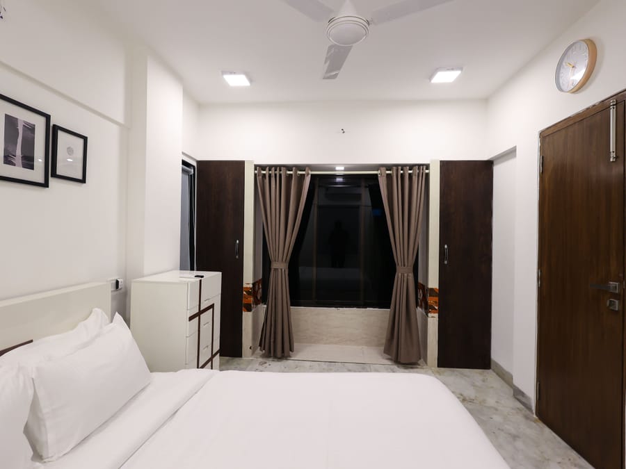 A Wabi-Sabi, Juhu bedroom with a white bed, 2 wall-mounted wardrobes flanking a curtained window, a white chest of drawers, and framed wall art.