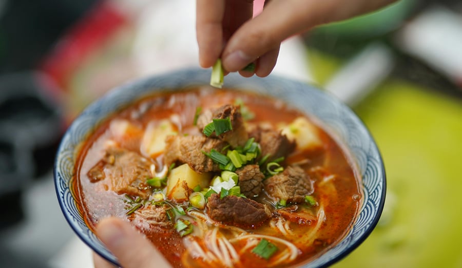 A bowl of noodle broth with vegetables and greens being finished with fresh garnish at The Garud Forest, Tadoba