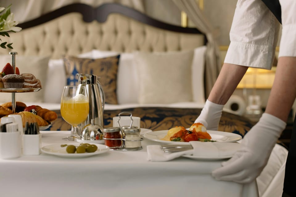 A white-gloved staff member serving a breakfast tray of eggs benedict, a tiered pastry stand, and fresh juice on a bedside table.