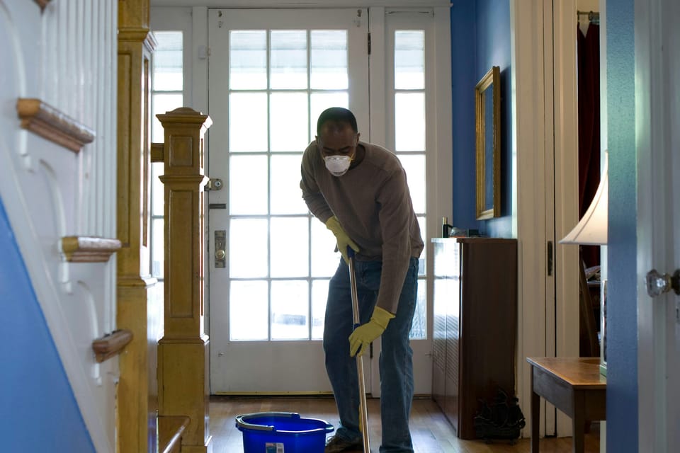 A person wearing a face mask and gloves mopping a wooden hallway floor beside a blue bucket near a staircase and a glass-panelled front door.
