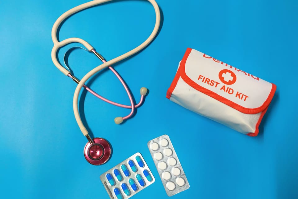 An overhead flat lay on a blue background featuring a pink stethoscope, a first aid kit pouch labelled with a cross, and 2 blister packs of pills.
