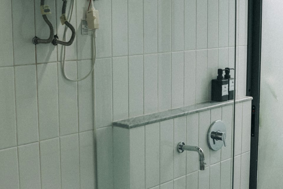 A tiled bathroom at Wabi-Sabi, Juhu with a wall-mounted water heater above the toilet, a glass shower partition, and wall-mounted taps.