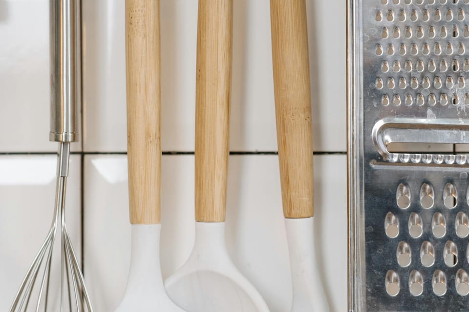 Kitchen utensils with wooden handles, including a whisk, a ladle, a spoon, and a spatula, hanging on hooks beside a metal grater against a tiled wall.