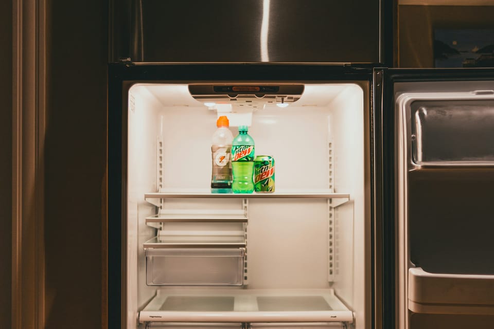 An open, mostly empty refrigerator with a few beverage bottles and a can on the top shelf, set into wooden kitchen cabinetry.