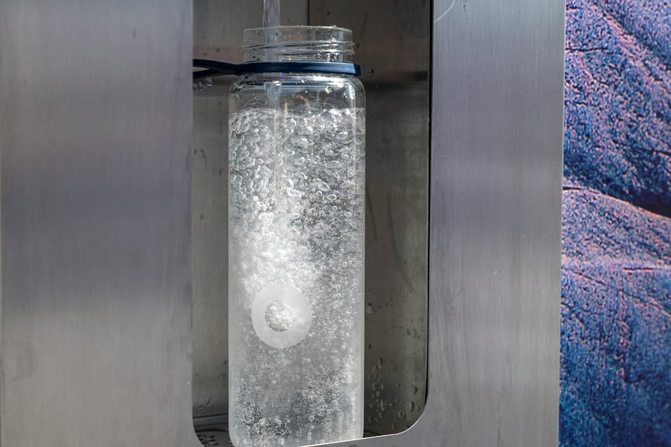 A glass bottle filling with water beneath the spout of a stainless steel Bluewater dispenser, with a hand pressing the button above.