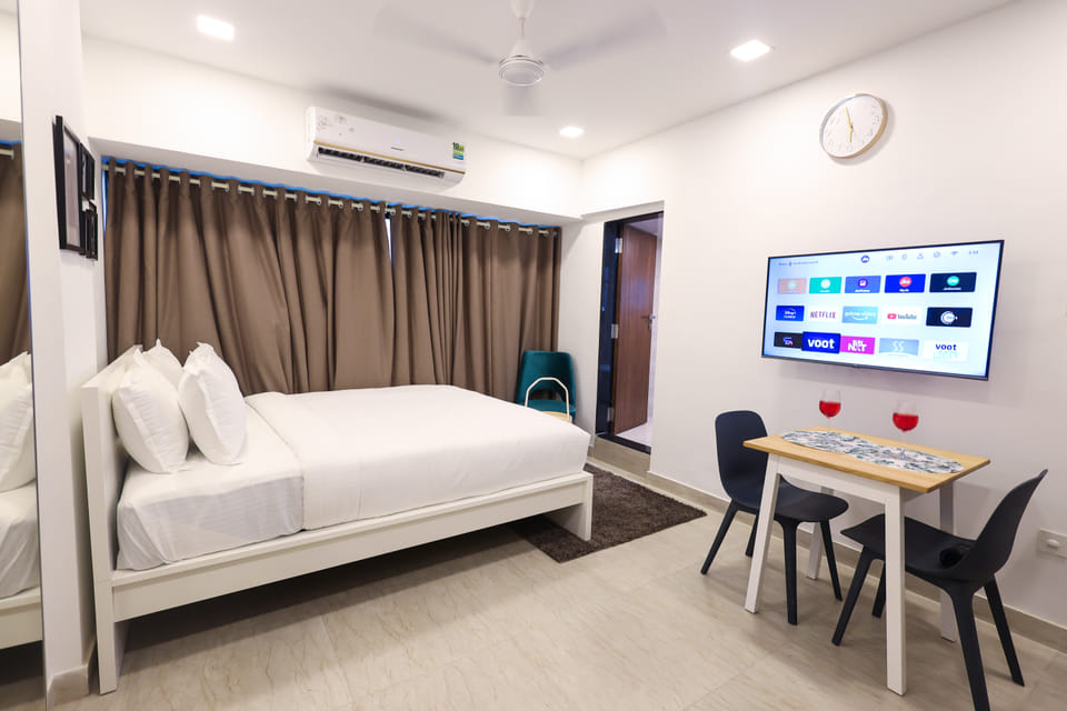 Bedroom at Wabi-Sabi, Juhu, with a bed, brown curtains, a wall-mounted AC, a smart TV showing streaming apps, and a dining table with 2 chairs and wine glasses.