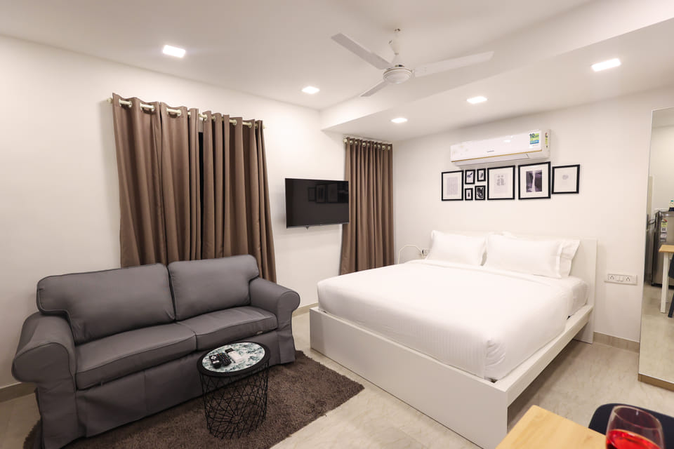 Bedroom at Wabi-Sabi, Juhu, with a white bed, a wall-mounted AC, a gallery wall of framed prints, a wall-mounted TV, and a 2-seater grey sofa.