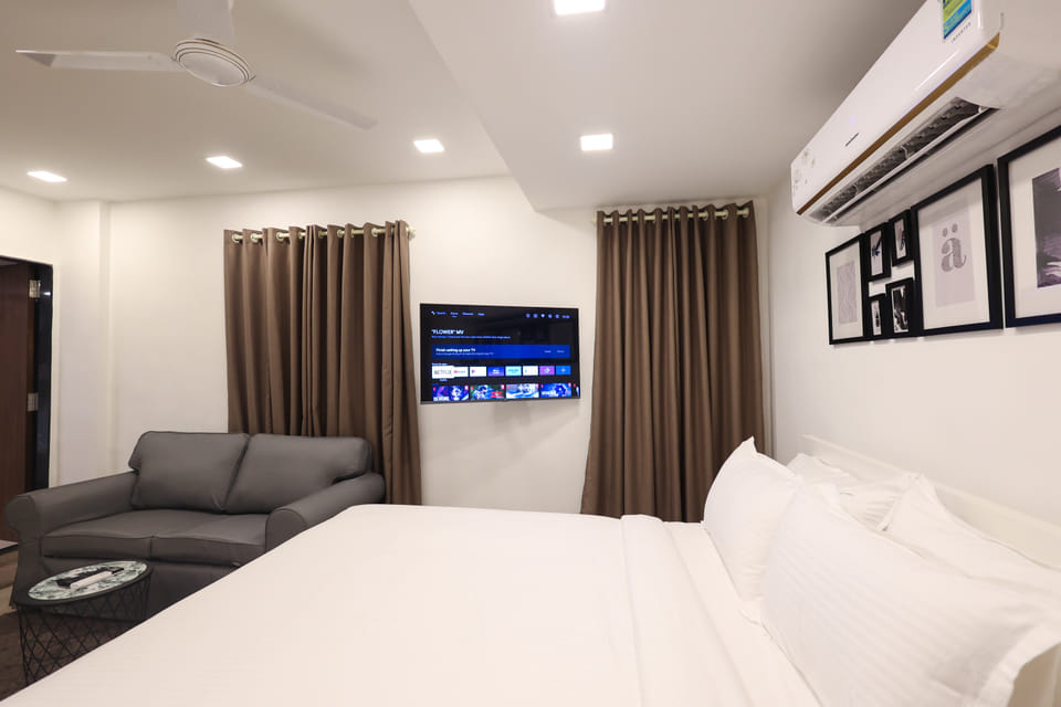 Bedroom at Wabi-Sabi, Juhu, with a neatly made bed, a wall-mounted TV, full-length curtains, a grey sofa, and a gallery wall of framed art.