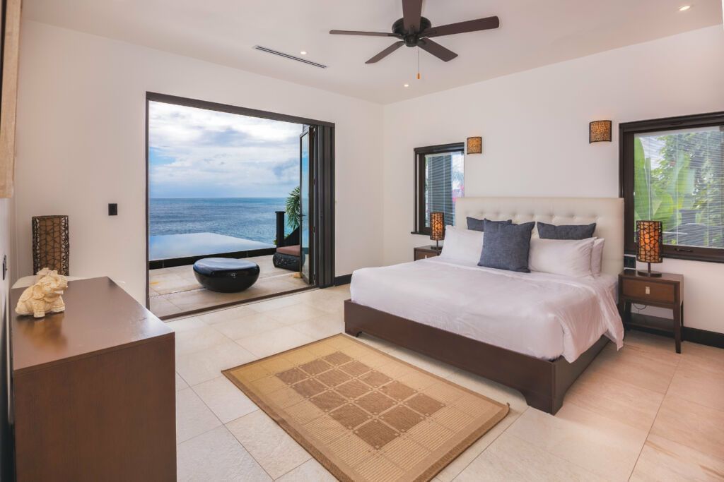 A modern ocean-view bedroom with a large bed, neutral tones, and glass doors opening to a patio and infinity pool overlooking the sea - Tamarind Hills Resort and Villas 