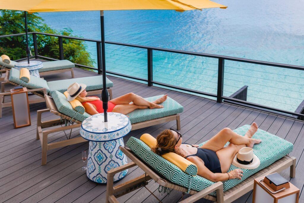 Two women lounging on sunbeds under a yellow umbrella on a wooden deck at Tamarind Hills Resort, Antigua, overlooking the turquoise Caribbean sea.