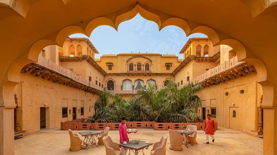 The central courtyard of Neemrana's Tijara Fort Palace, viewed through an ornate archway, showcases seating areas and lush greenery within the historic structure.