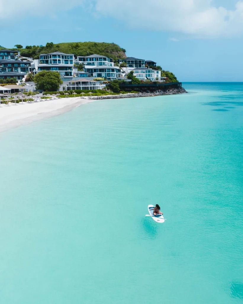 A lone paddleboarder on calm turquoise waters near a hillside resort with lush greenery - Tamarind Hills Resort and Villas - Antigua