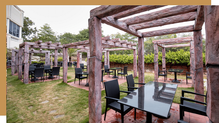 Outdoor Eden Garden dining area with tables and chairs arranged under wooden pergolas on a grassy lawn, surrounded by greenery and hedges, with a building visible in the background.