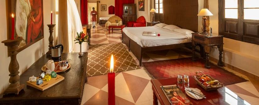 A richly decorated bedroom at Neemrana's Tijara Fort Palace includes a bed, seating area with red accents, artwork, and a tea/coffee setup.