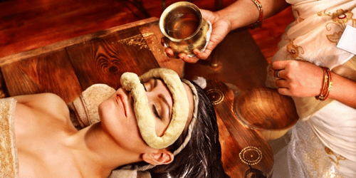 A person lying on a surface while a practitioner pours liquid from a vessel onto their forehead