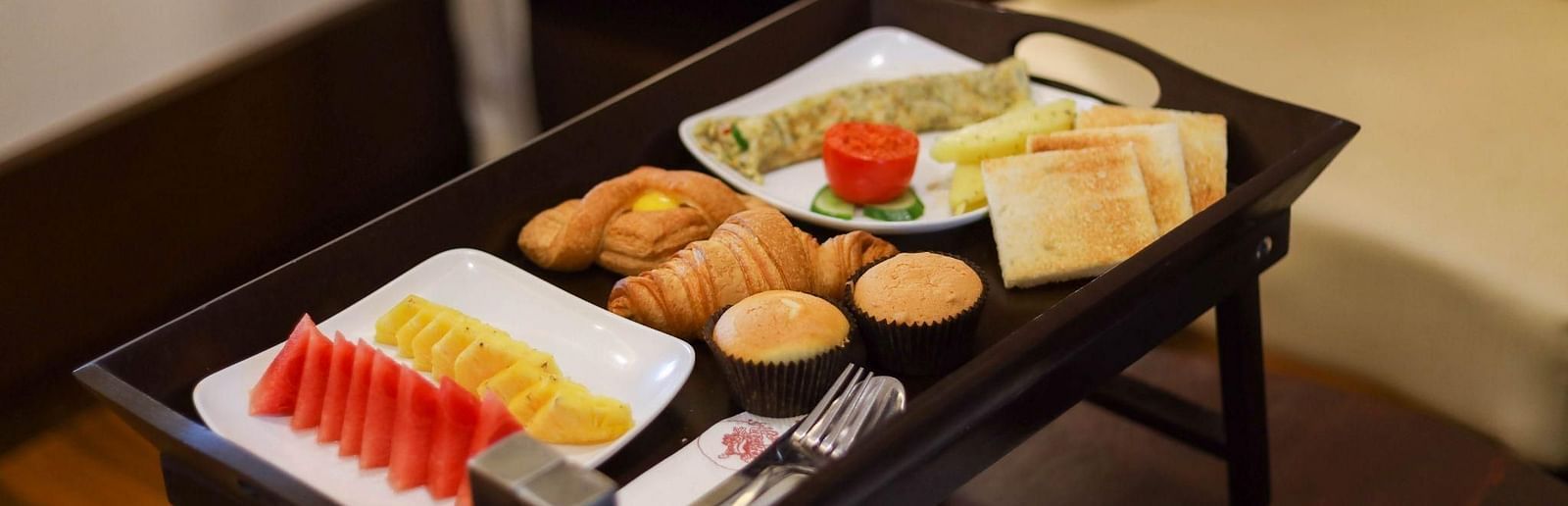 Room service breakfast tray with toast, muffins, croissants, omelette, fresh fruit, and cutlery set, placed on a table iin Avana Resort