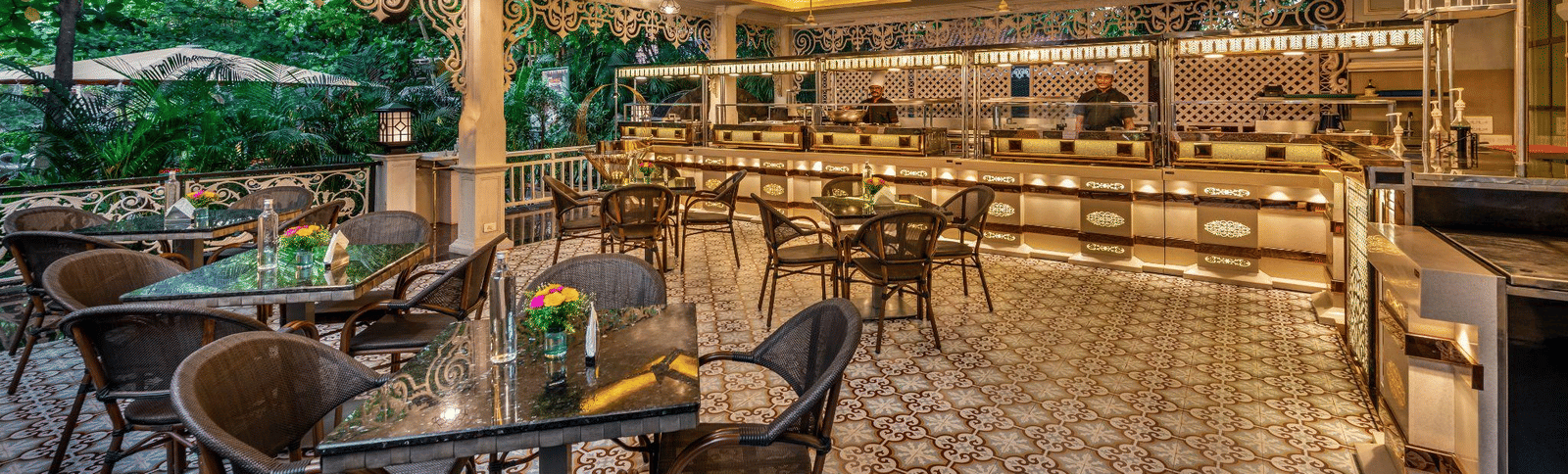 Elegant dining space with a golden ceiling, decorative lighting, a long buffet setup with chafing dishes, and black wicker chairs.