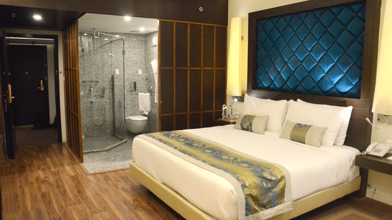 A room with a neatly made bed and bedside tables, warm lighting, and a doorway leading to a modern washroom at Clarks Avadh, Lucknow | Rooms with Gomti River View.
