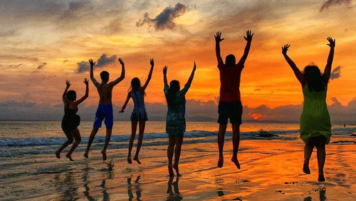 A family standing on a beach at sunset, jumping with their hands raised, with their reflections visible on the wet sand and the sky filled with clouds.