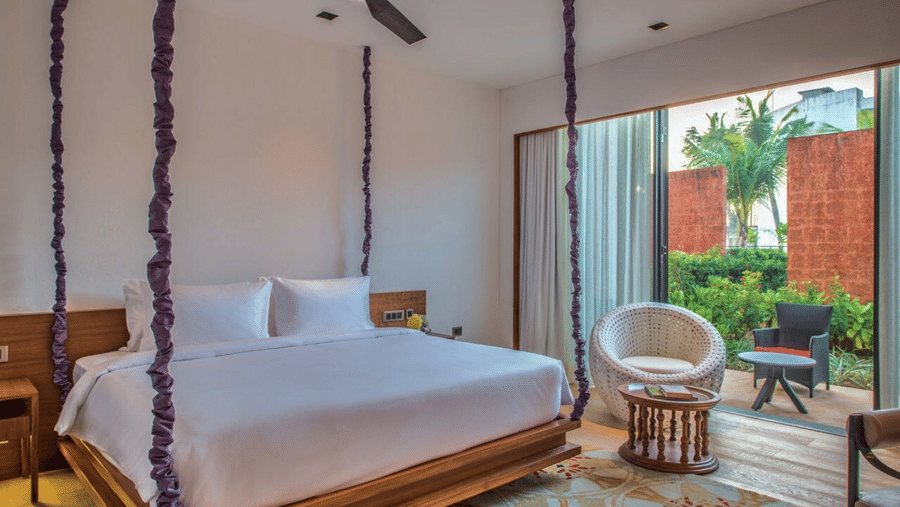 The Deluxe Patio King Room with a large wooden bed hanging from the ceiling on purple rope vines, and an armchair at Azaya Beach Resort, Goa.
